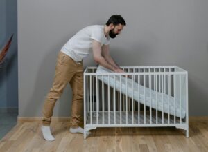 Young man adjusting a crib mattress in a modern, minimalist nursery with hardwood floors.
